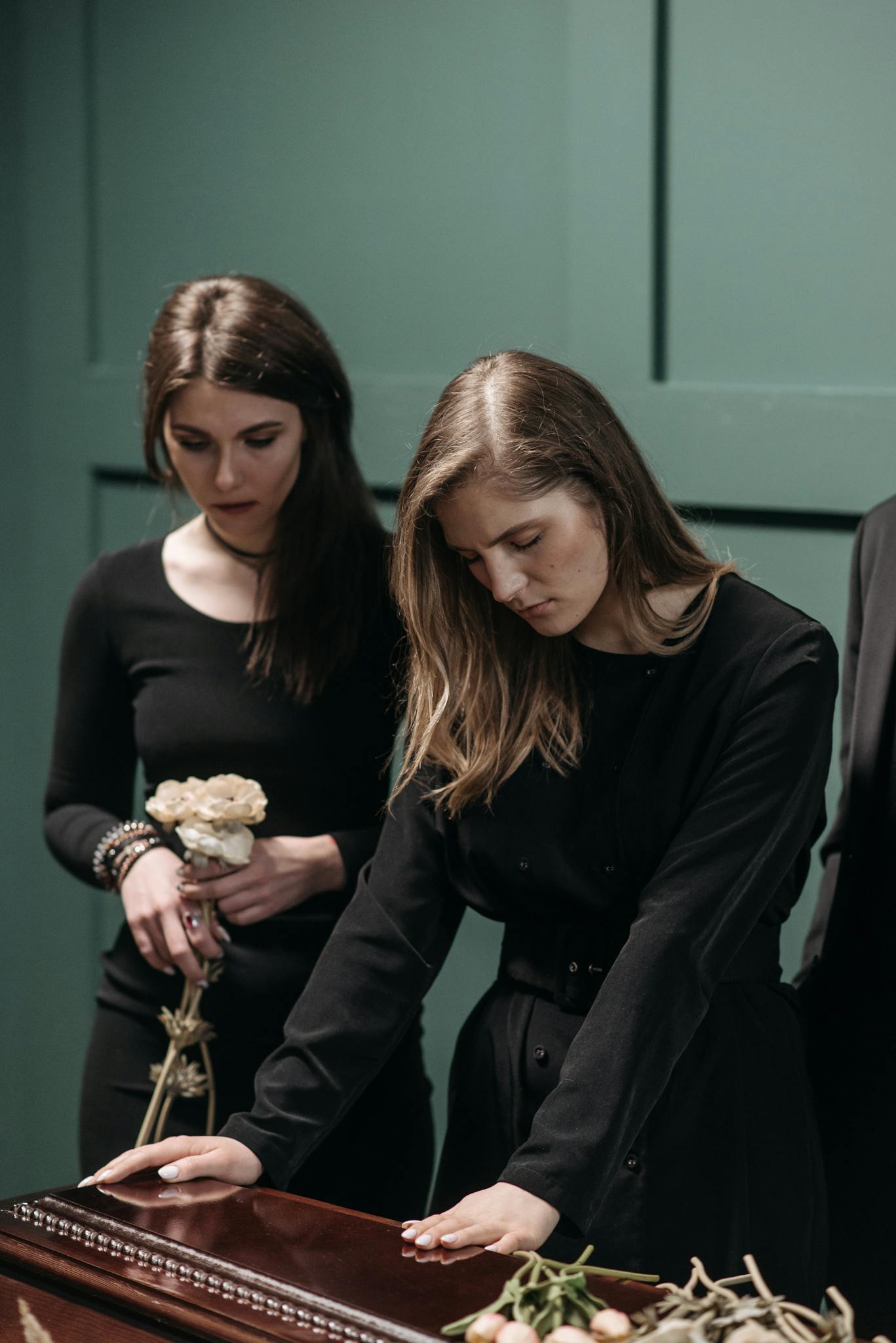 Two women expressing grief by a coffin, holding flowers in a solemn funeral setting.